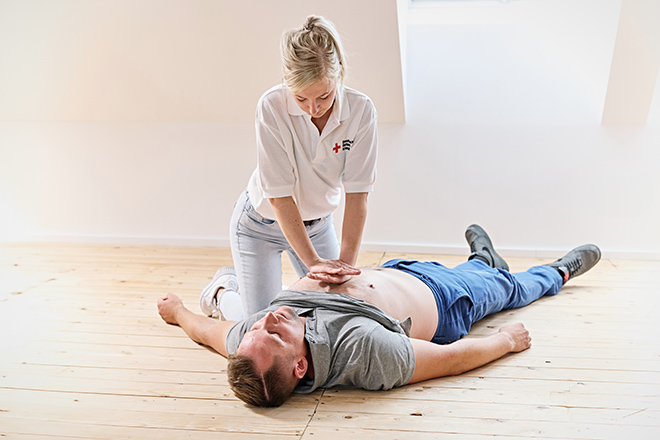 Auf dem Boden liegt ein Mann auf dem Rücken, die Arme seitlich ausgebreitet; darüber beugt sich eine Frau, die eine Herz-Druck-Massage demonstriert